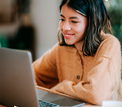 mujer sonriendo viendo una laptop, esta sentada en una mesa, lleva suéter beige y tiene cabello negro con destellos amarillos.
